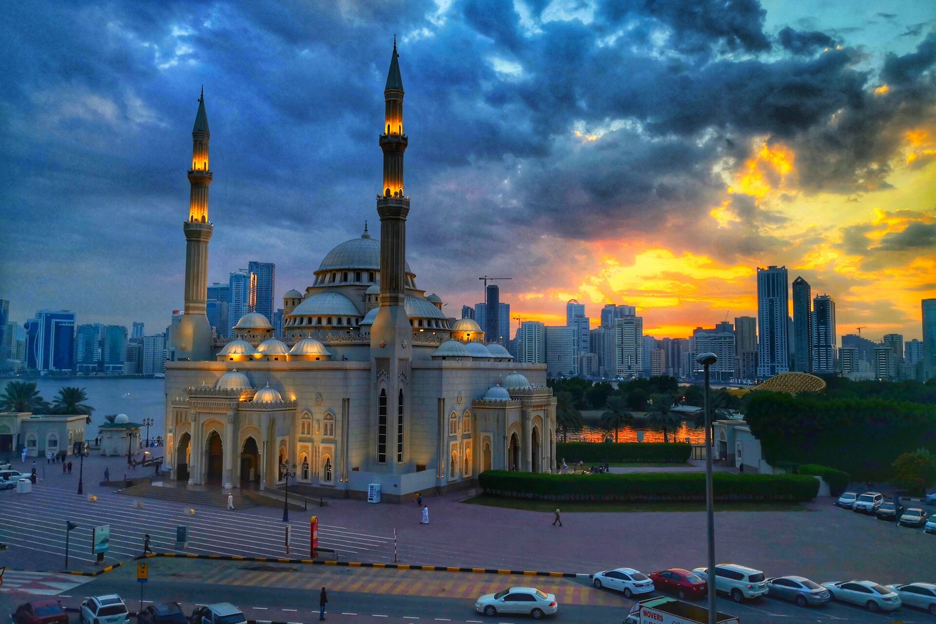 Al Noor Mosque lit at sunset beside Khalid Lagoon, with the Sharjah skyline beyond