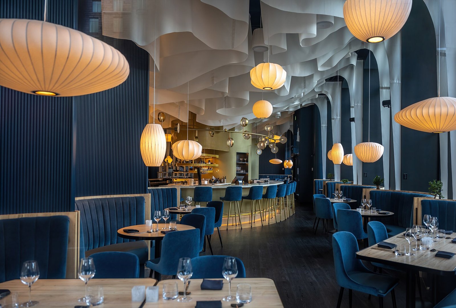 An elegant dining room with deep blue banquettes, set tables and glowing paper lanterns beneath a sculpted ceiling