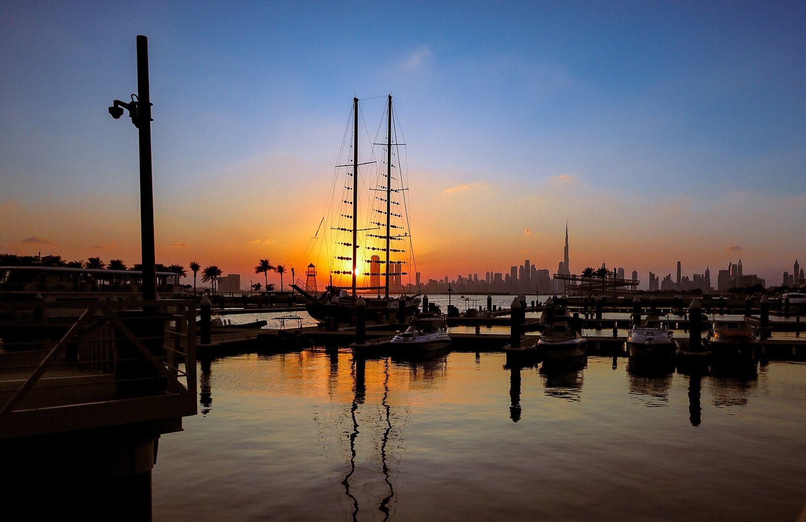 The masts of a moored sailing ship silhouetted against a golden sunset over Dubai Creek Harbour, with the Dubai skyline beyond.