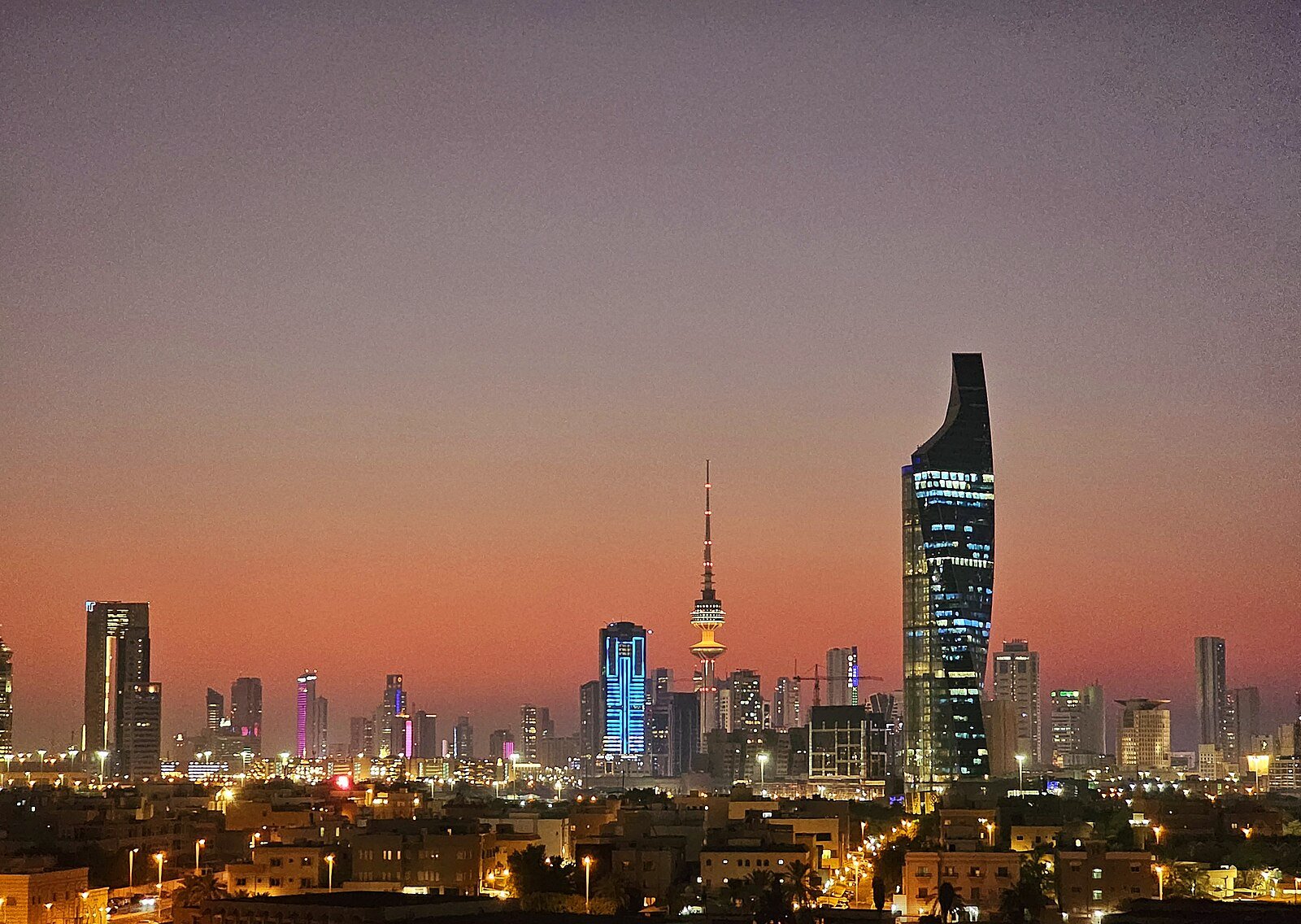 The Kuwait City skyline at dusk, with the Al Hamra Tower and Liberation Tower lit against a rose-colored sky.