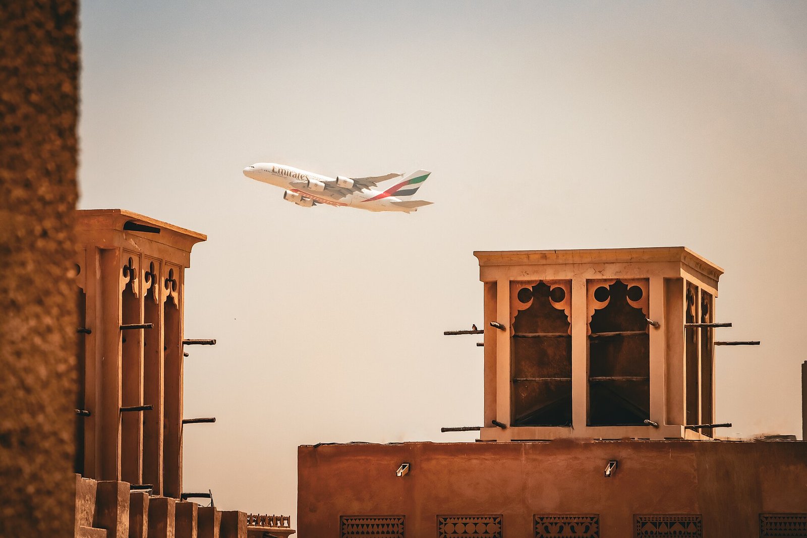 An Emirates A380 passes over the wind towers of Dubai's Al Fahidi Historical Neighbourhood in warm evening light.