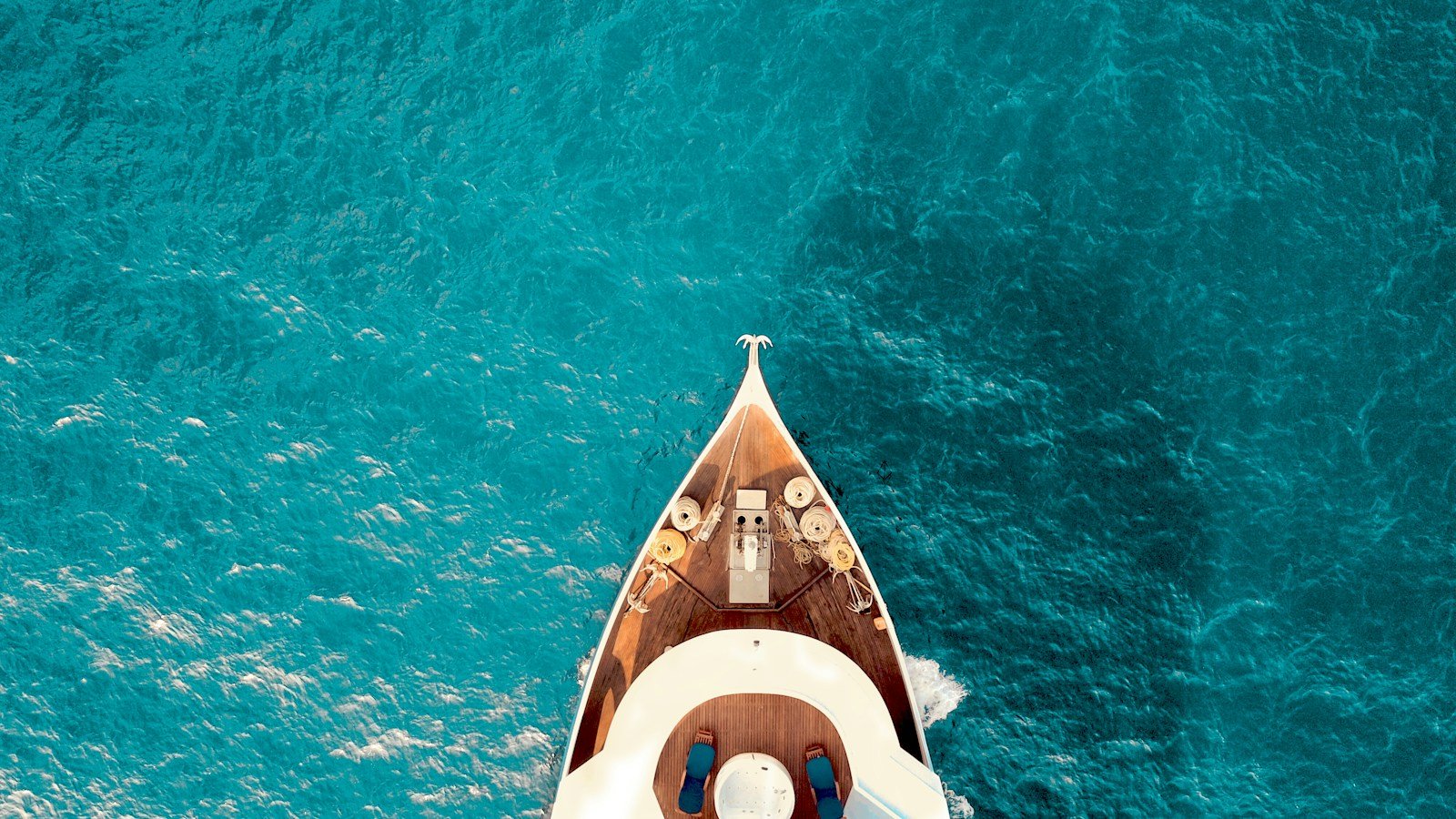 Looking down on a yacht's teak foredeck and sun loungers as it cuts through turquoise water.