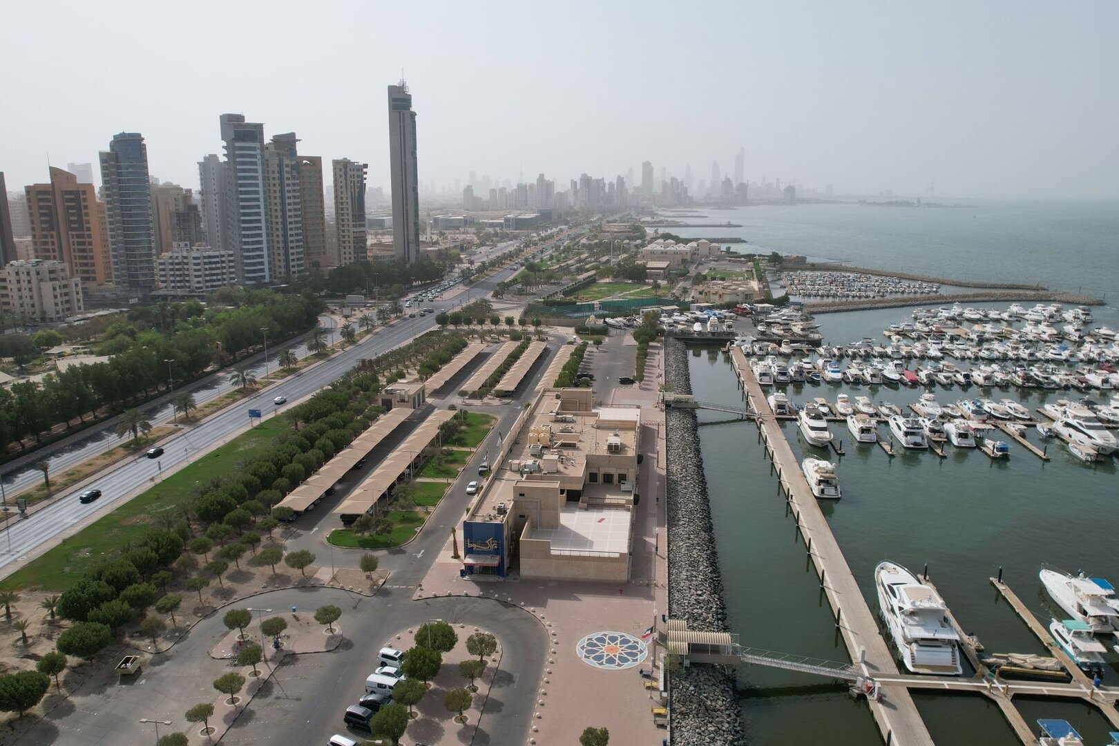 A marina full of yachts beside the Gulf Road, with the towers of Kuwait City beyond