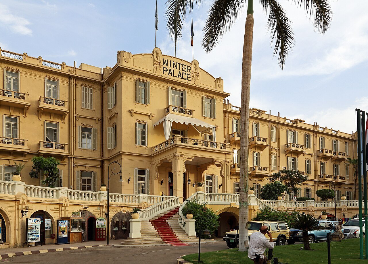 The ochre facade and grand red-carpeted staircase of the Winter Palace hotel in Luxor, framed by palms.