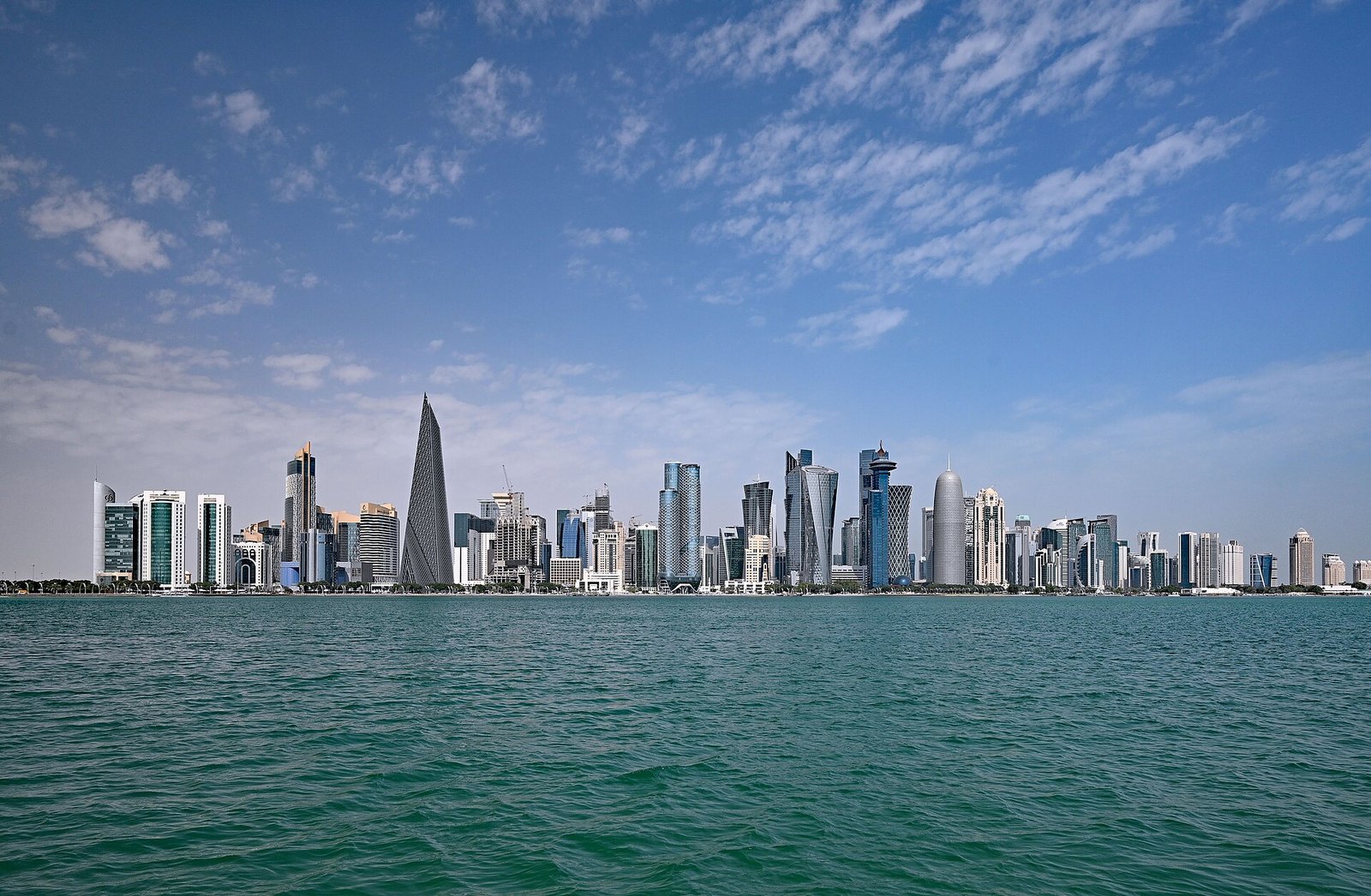 The towers of Doha's West Bay rising above the turquoise water of Doha Bay under a cloud-streaked sky.