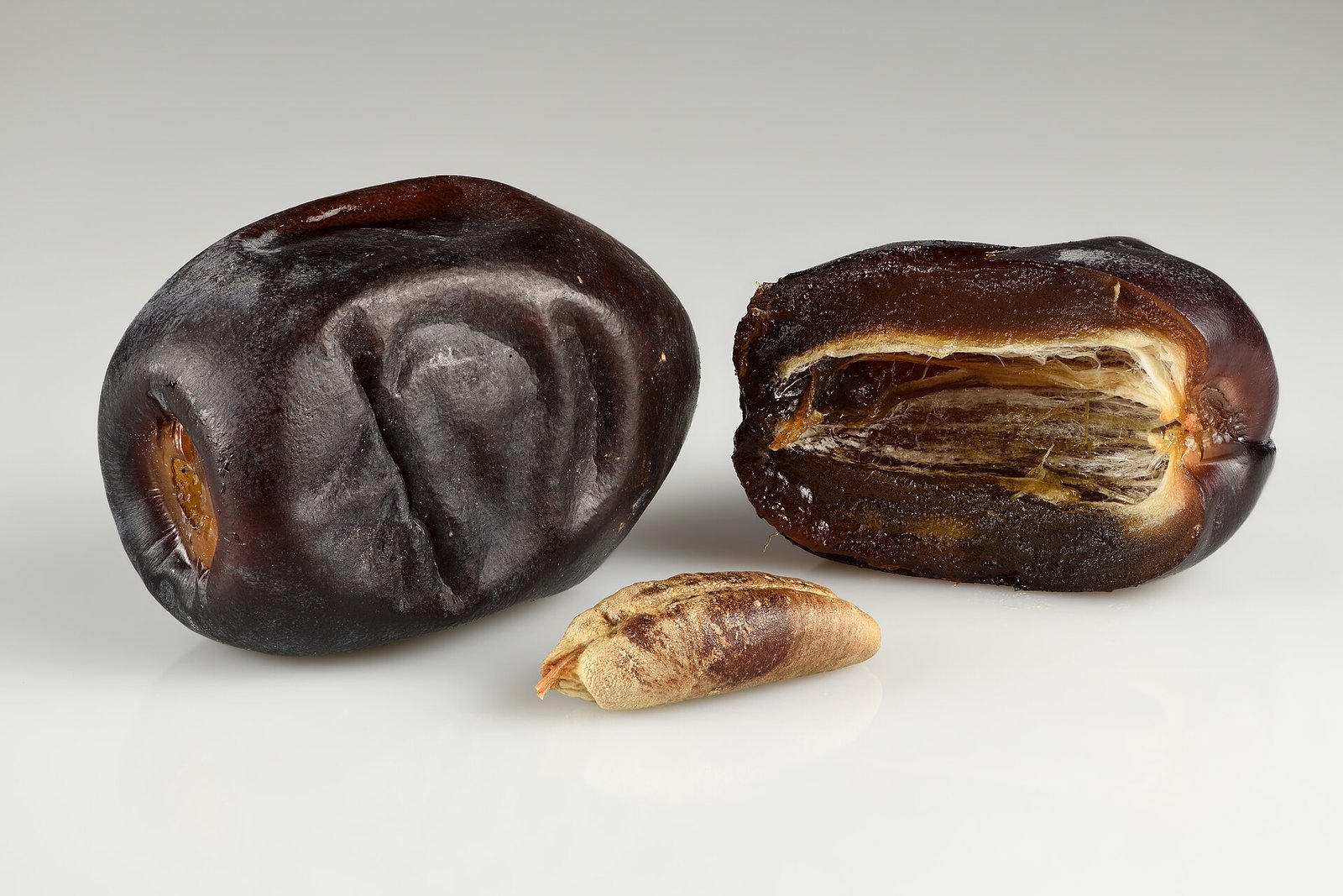 Glossy Mazafati dates, one whole and one halved beside its seed, on a pale background.