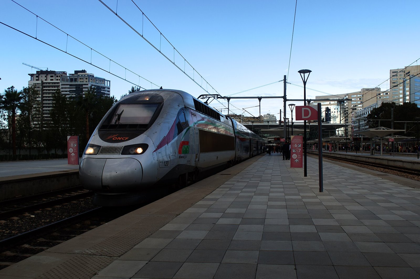 A silver Al Boraq high-speed train waiting at the platform of Tanger-Ville station under a blue sky.