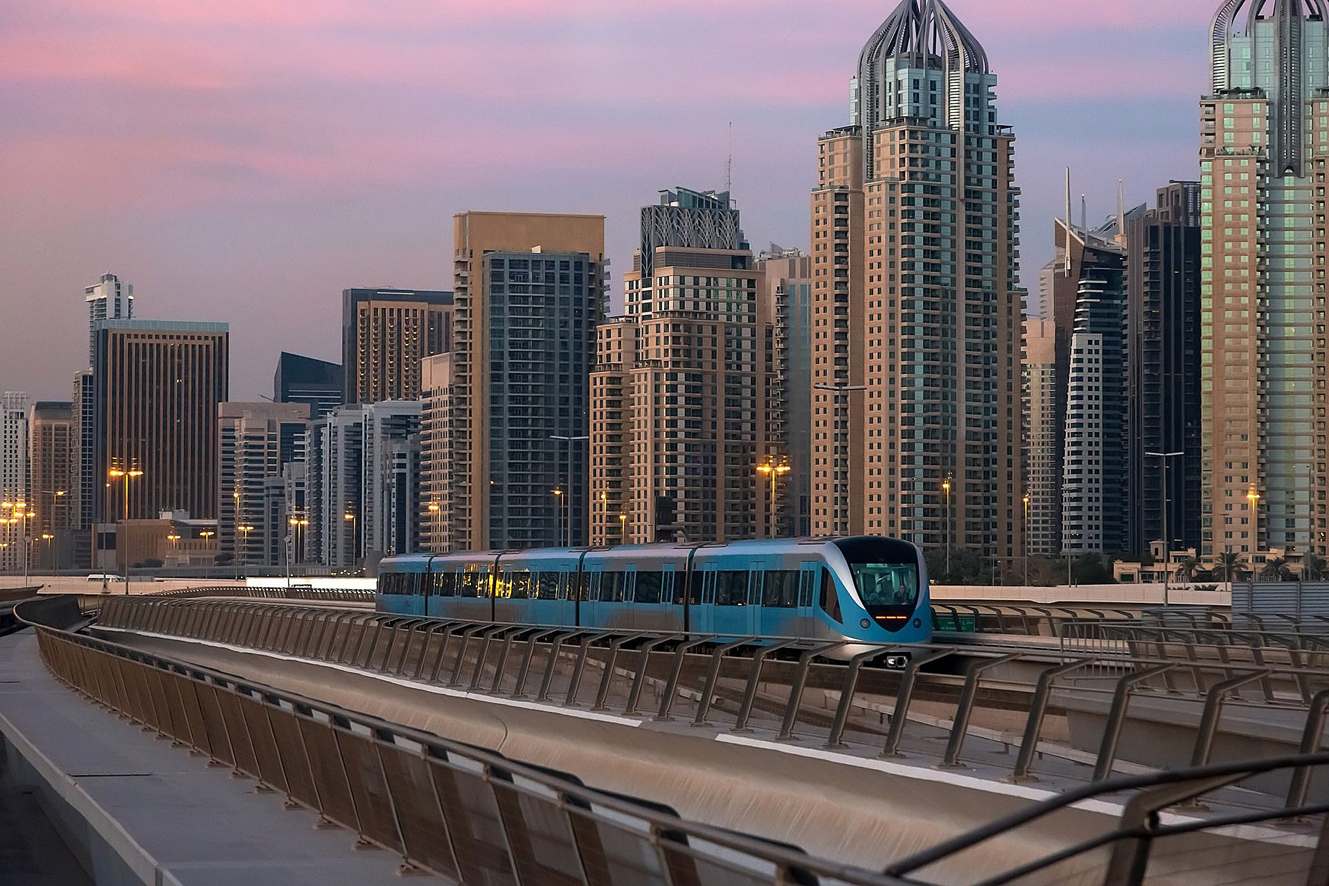 A blue Dubai Metro train gliding along its elevated track at dusk, the towers of Dubai Marina rising behind