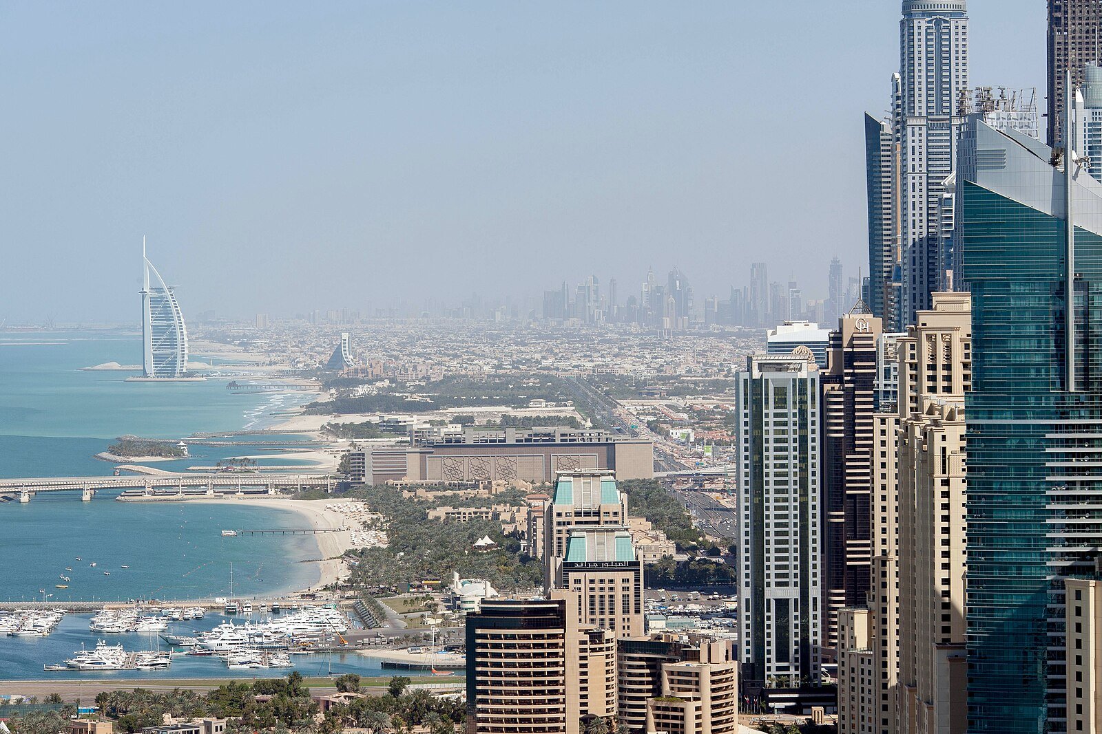 The Dubai coastline curving toward the sail of the Burj Al Arab, with marina towers and beaches below.