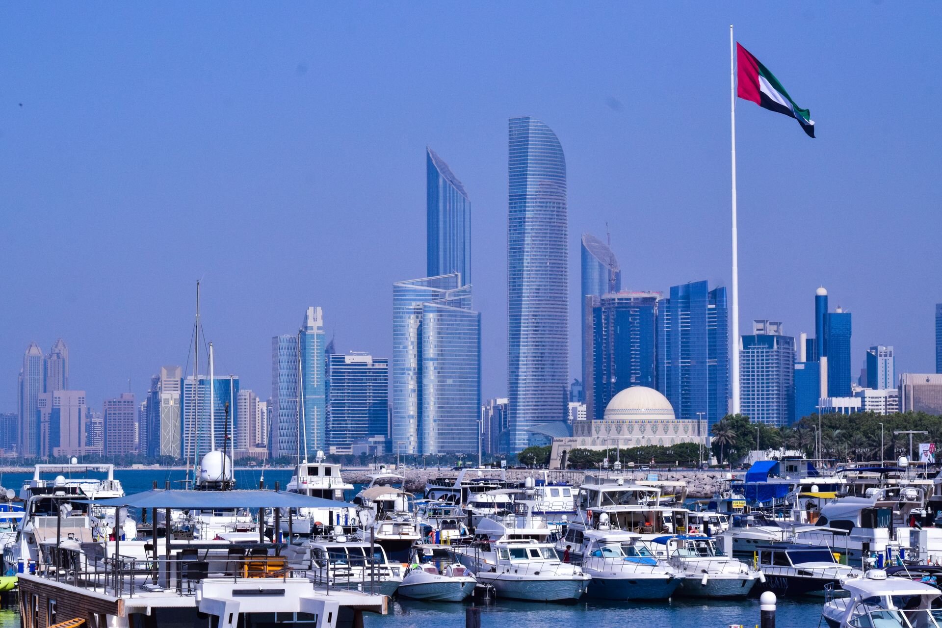 The towers of Abu Dhabi beyond a Corniche marina, under the UAE flag