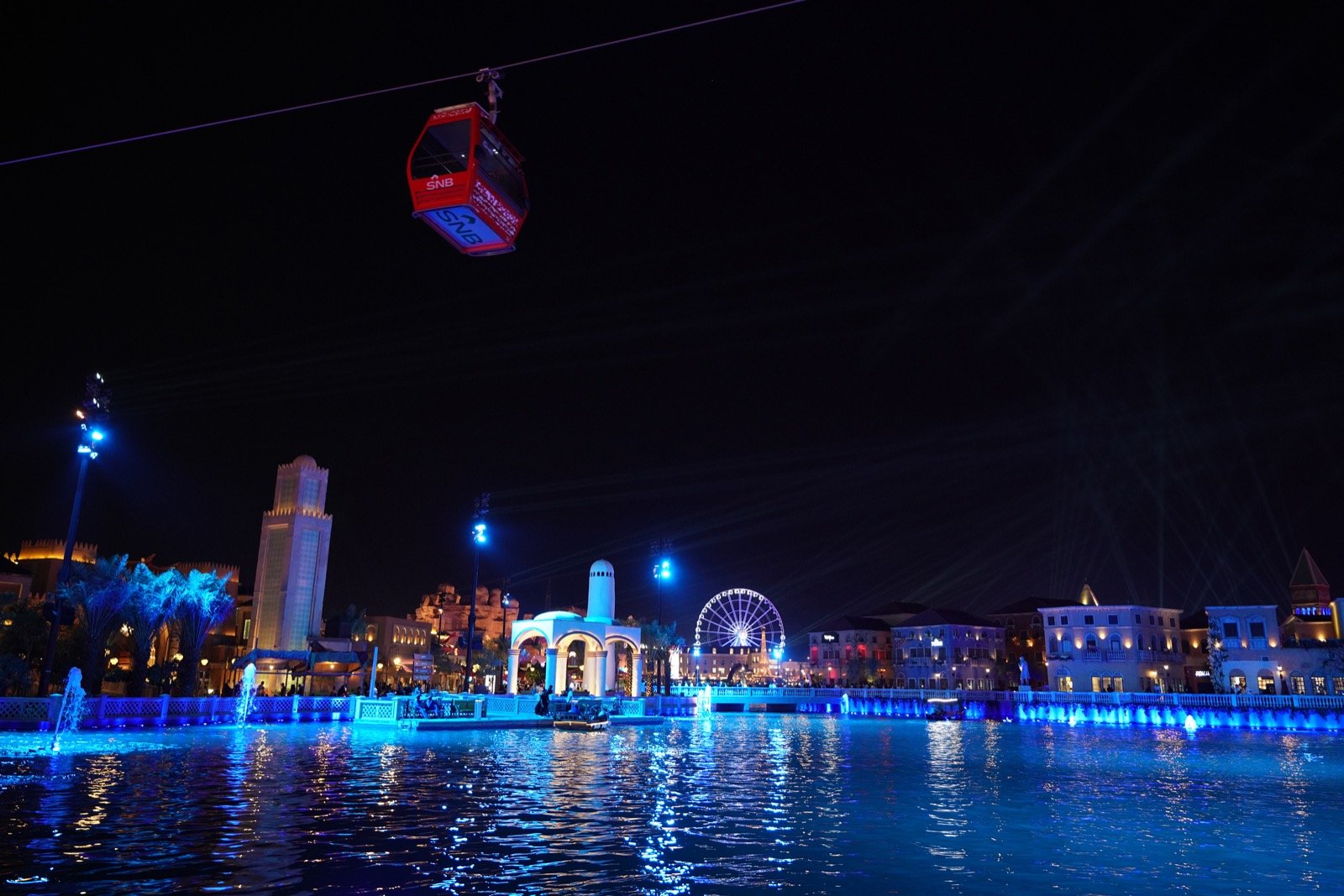 Boulevard Riyadh City lit in blue at night, with a cable car gliding above its lagoon and pavilions.