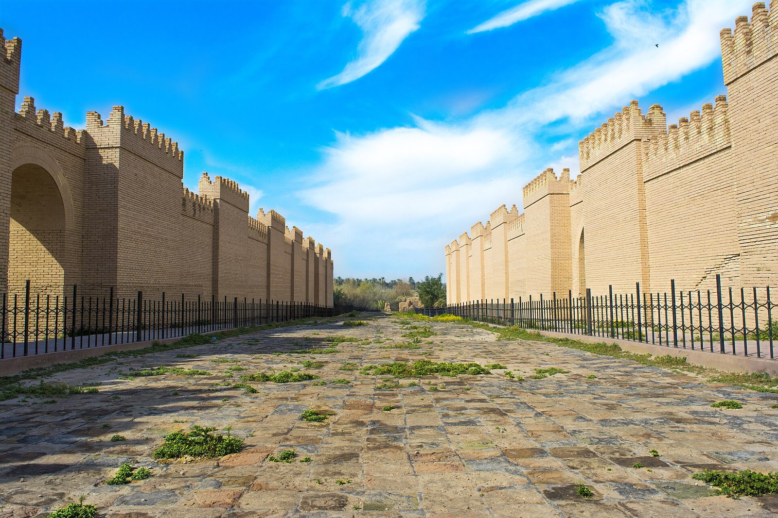 The reconstructed crenellated walls of ancient Babylon lining a paved processional street under a bright blue sky.