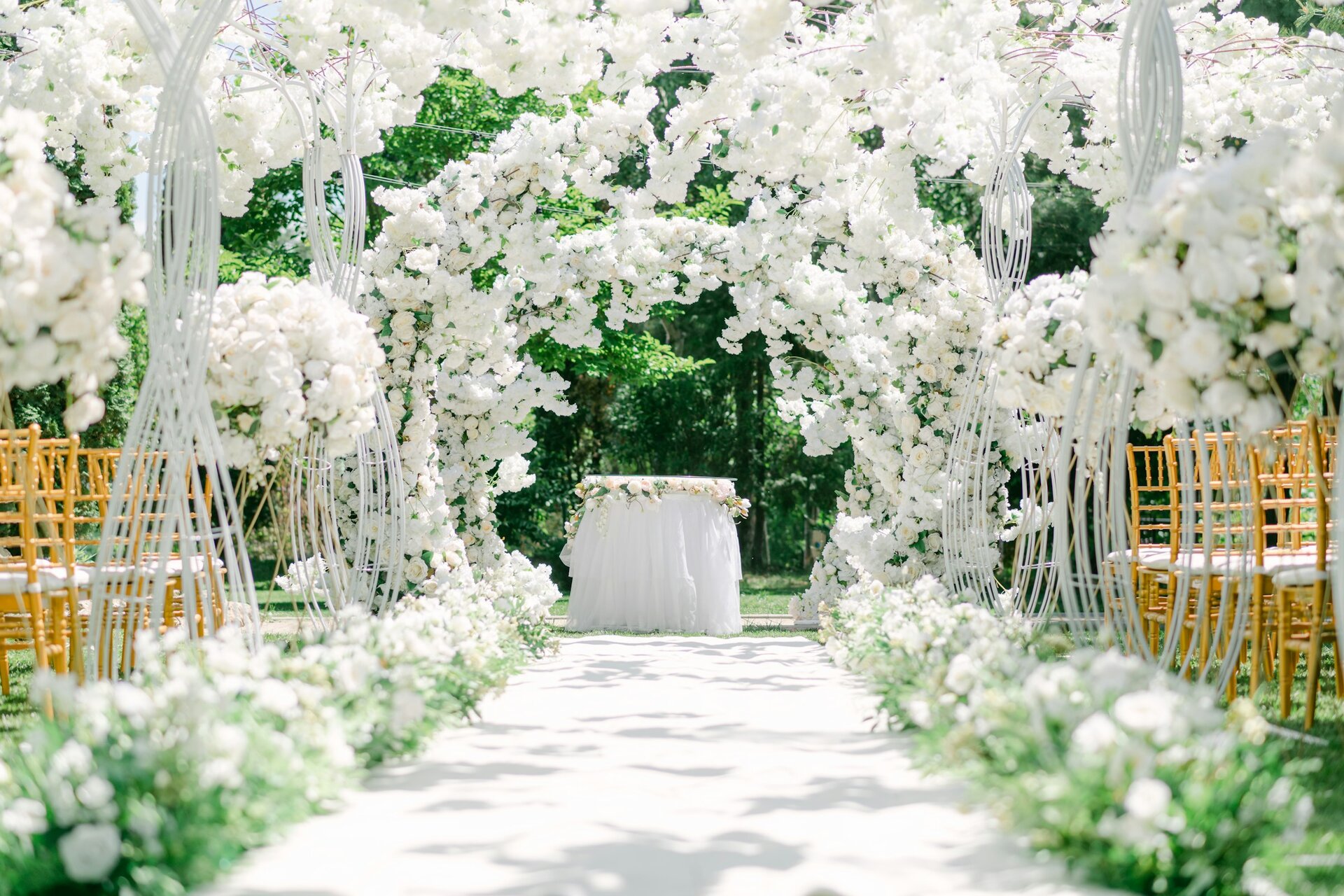 A wedding aisle beneath arches of white flowers, lined with gold chairs