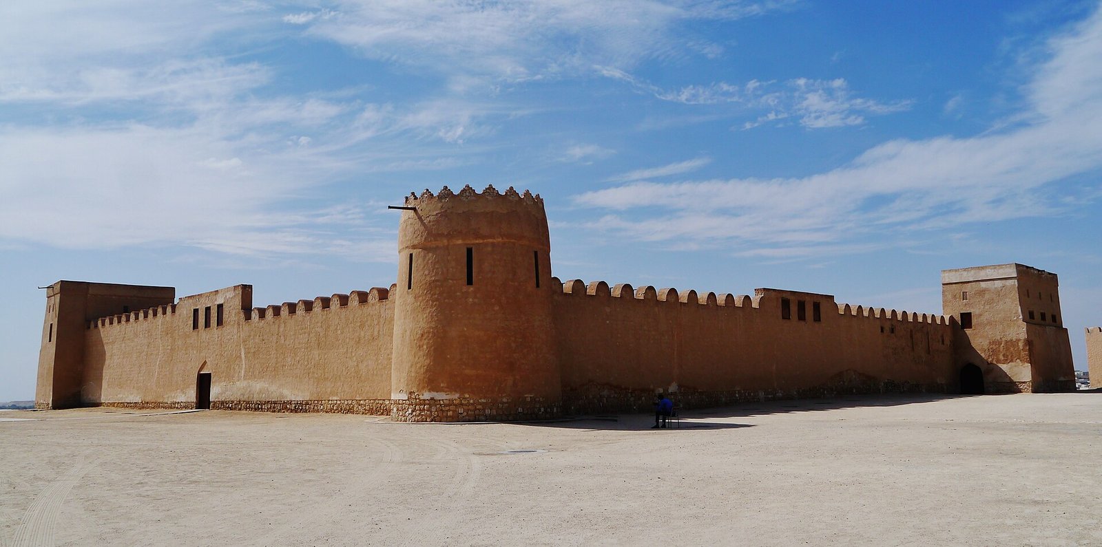 The sand-colored walls and round tower of Riffa Fort, Sheikh Salman bin Ahmed Al Fateh Fort, in Bahrain under a blue sky.