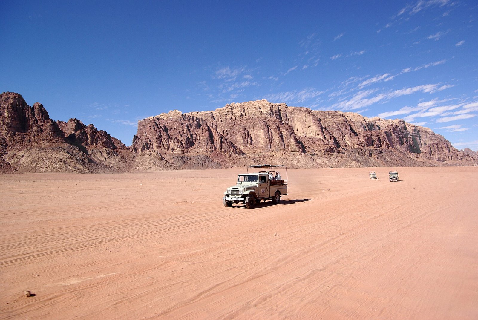 A four-by-four crossing the red sands of Wadi Rum below towering sandstone cliffs.