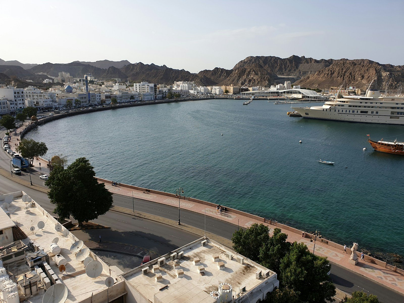 The curving Muttrah Corniche and harbour of Muscat, with a royal yacht and a wooden dhow beneath the Hajar Mountains.