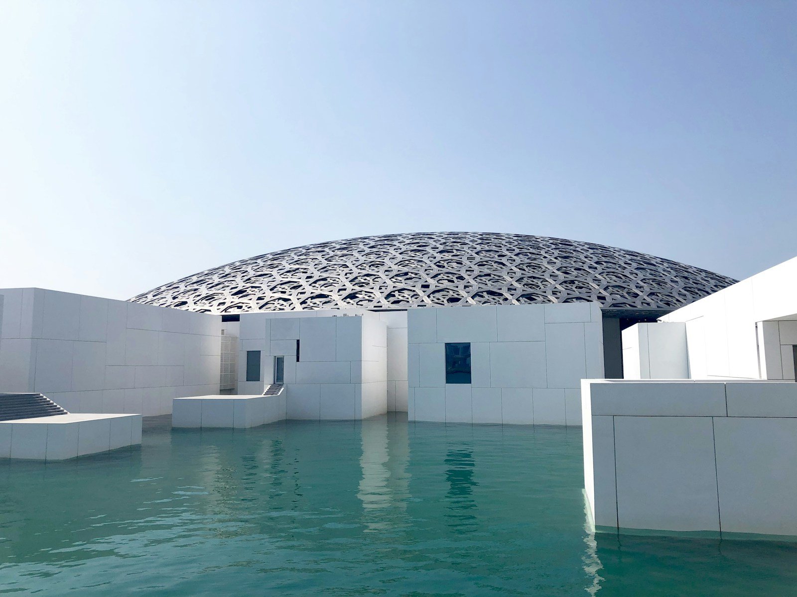The latticed silver dome of the Louvre Abu Dhabi floating above white galleries and still sea water on Saadiyat Island.