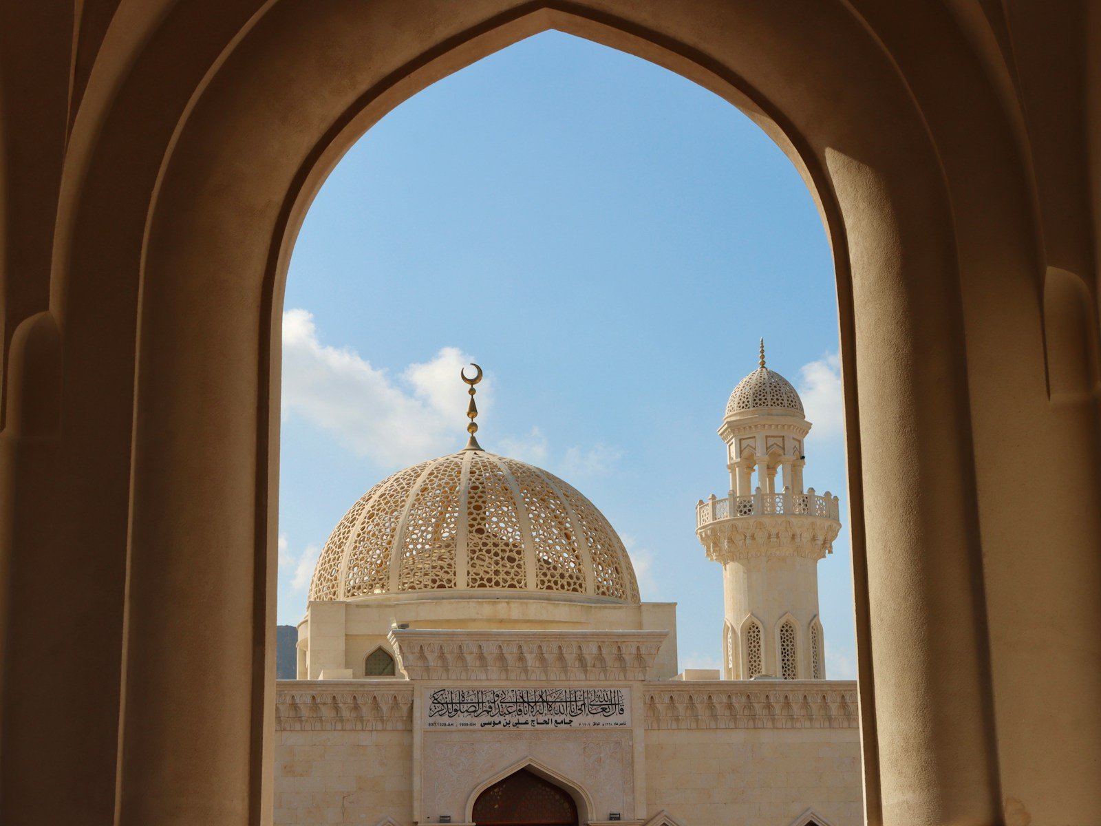 The latticed golden dome and a minaret of the Sultan Qaboos Grand Mosque framed by a pointed sandstone arch, Muscat.