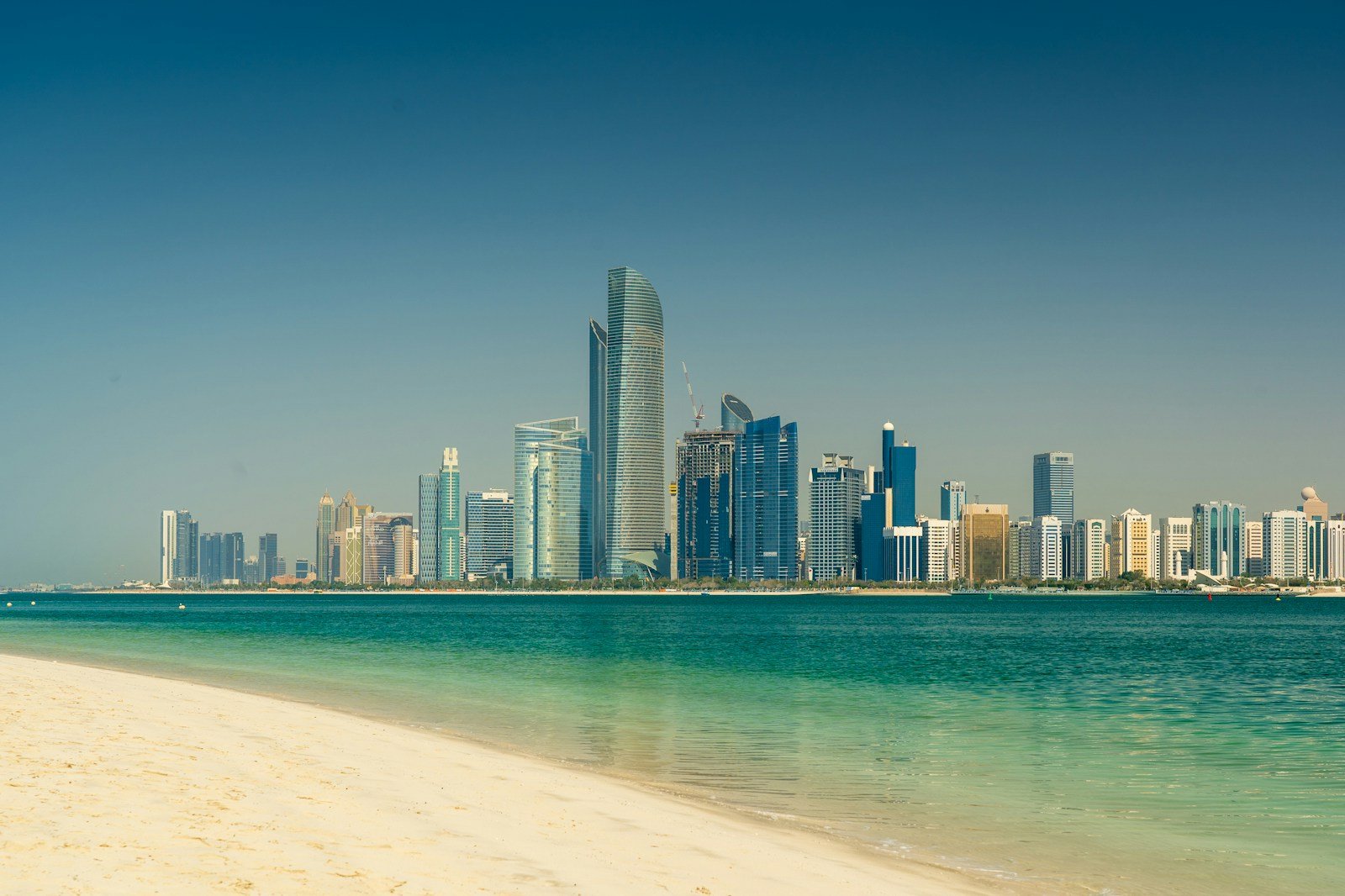 The towers of the Abu Dhabi Corniche rising across clear turquoise water from a white-sand beach.