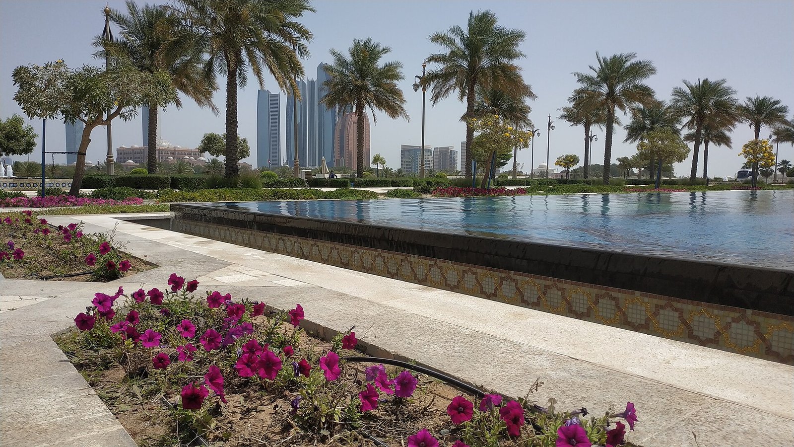 A long reflecting pool, flowerbeds and date palms in the gardens of Qasr Al Watan, with the towers of Abu Dhabi beyond.
