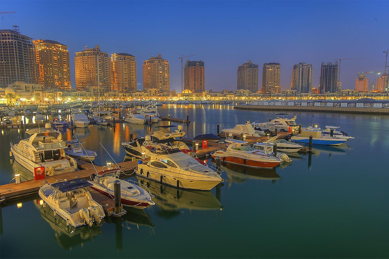 Yachts moored at The Pearl marina in Doha at blue hour, the waterfront towers lit behind them.