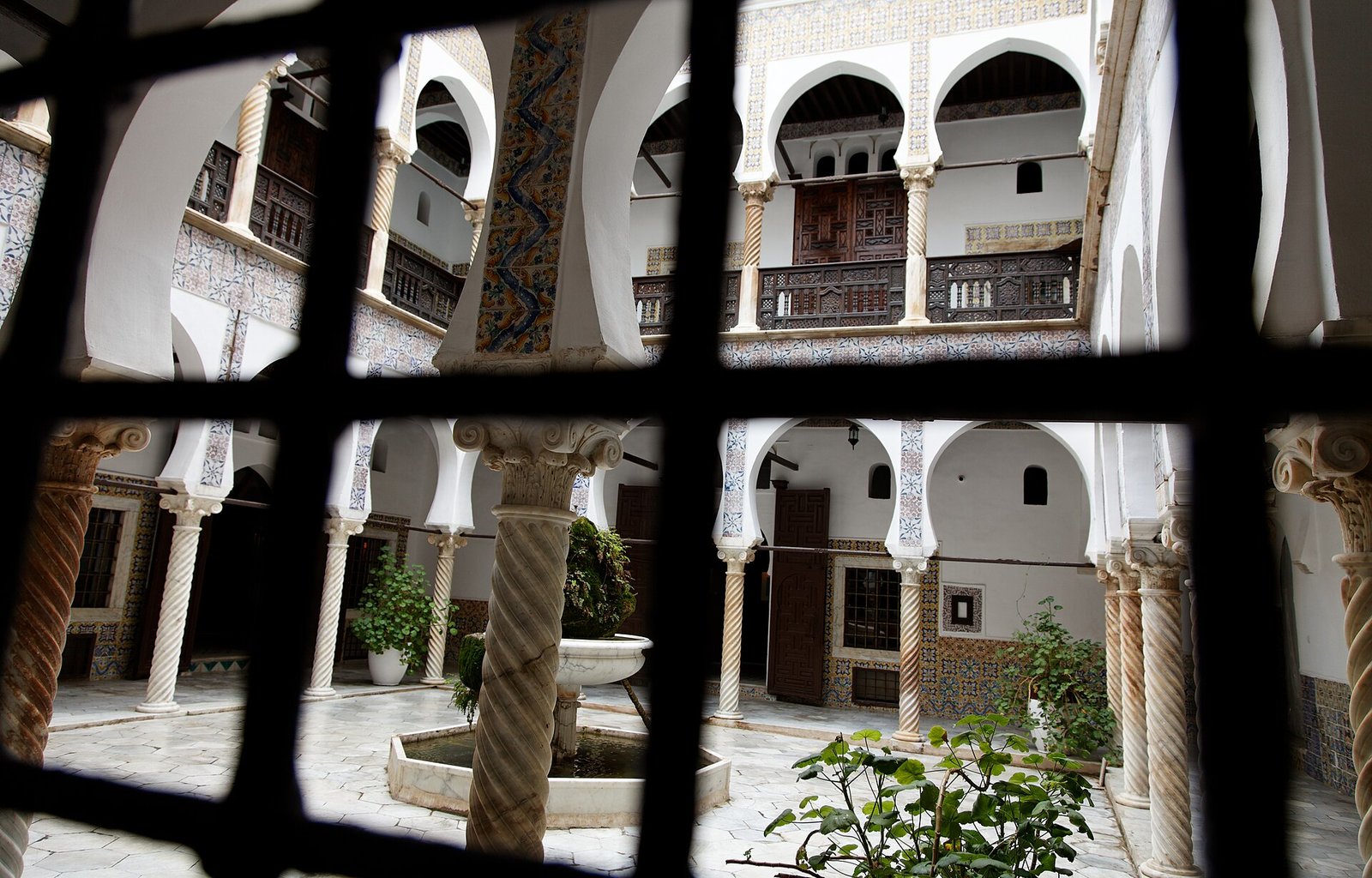 The carved columns and horseshoe arches of an Ottoman-era courtyard house in the Casbah of Algiers, glimpsed through a wrought-iron window grille.