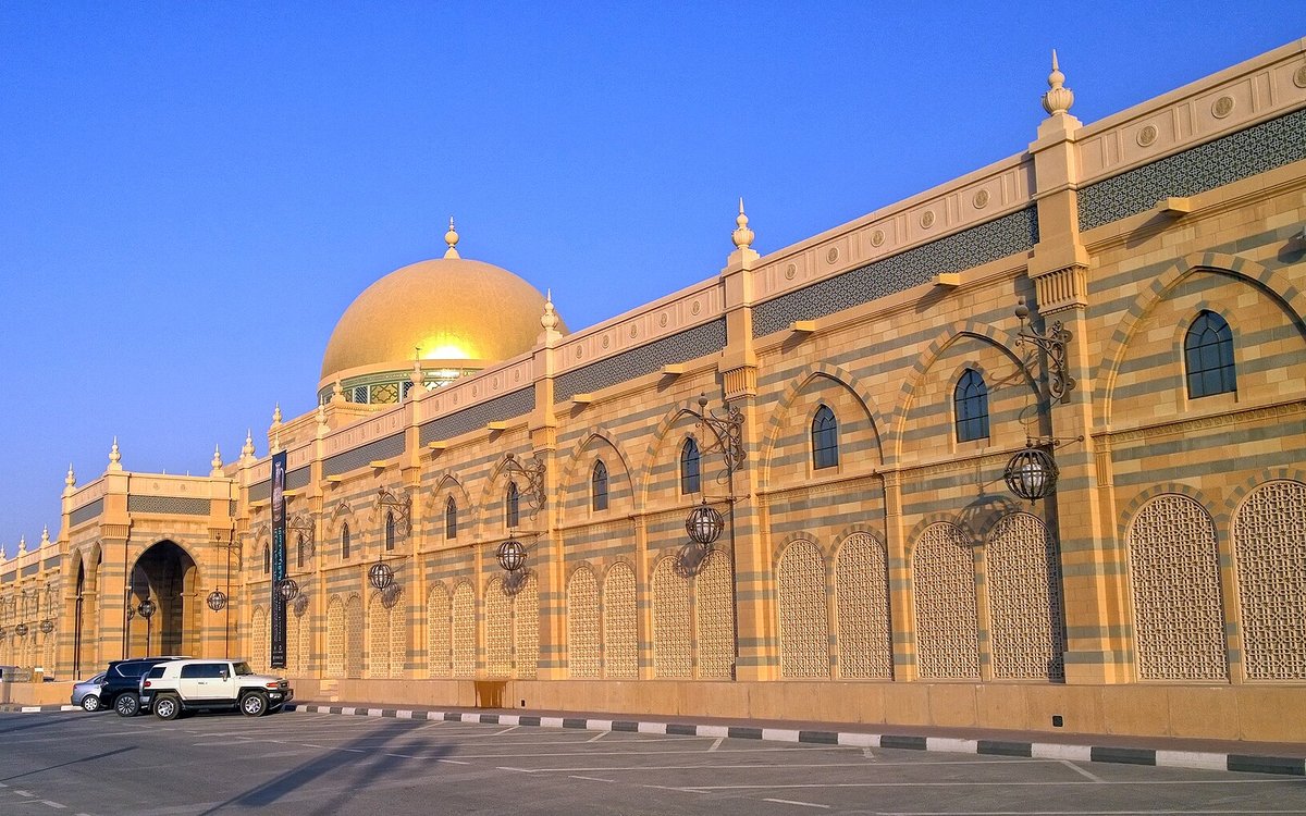 The golden dome and striped stone arcades of the Sharjah Museum of Islamic Civilization shine under a deep blue sky.
