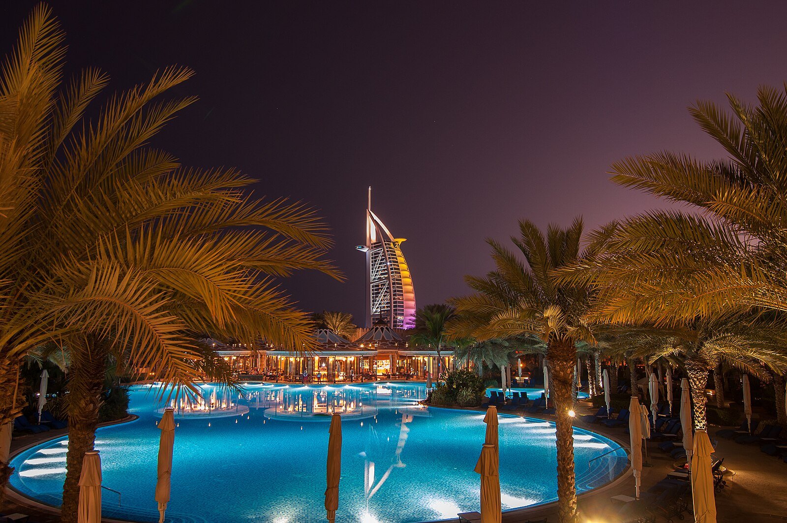 The illuminated sail of the Burj Al Arab rising beyond palm trees and a softly lit resort pool at dusk in Dubai