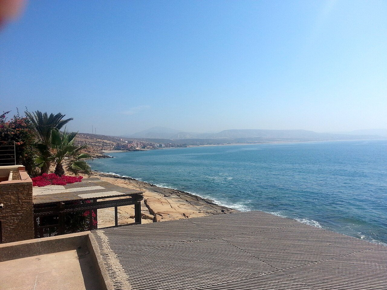 A palm-shaded terrace above the Atlantic coastline at Taghazout, Morocco, on a bright clear day.