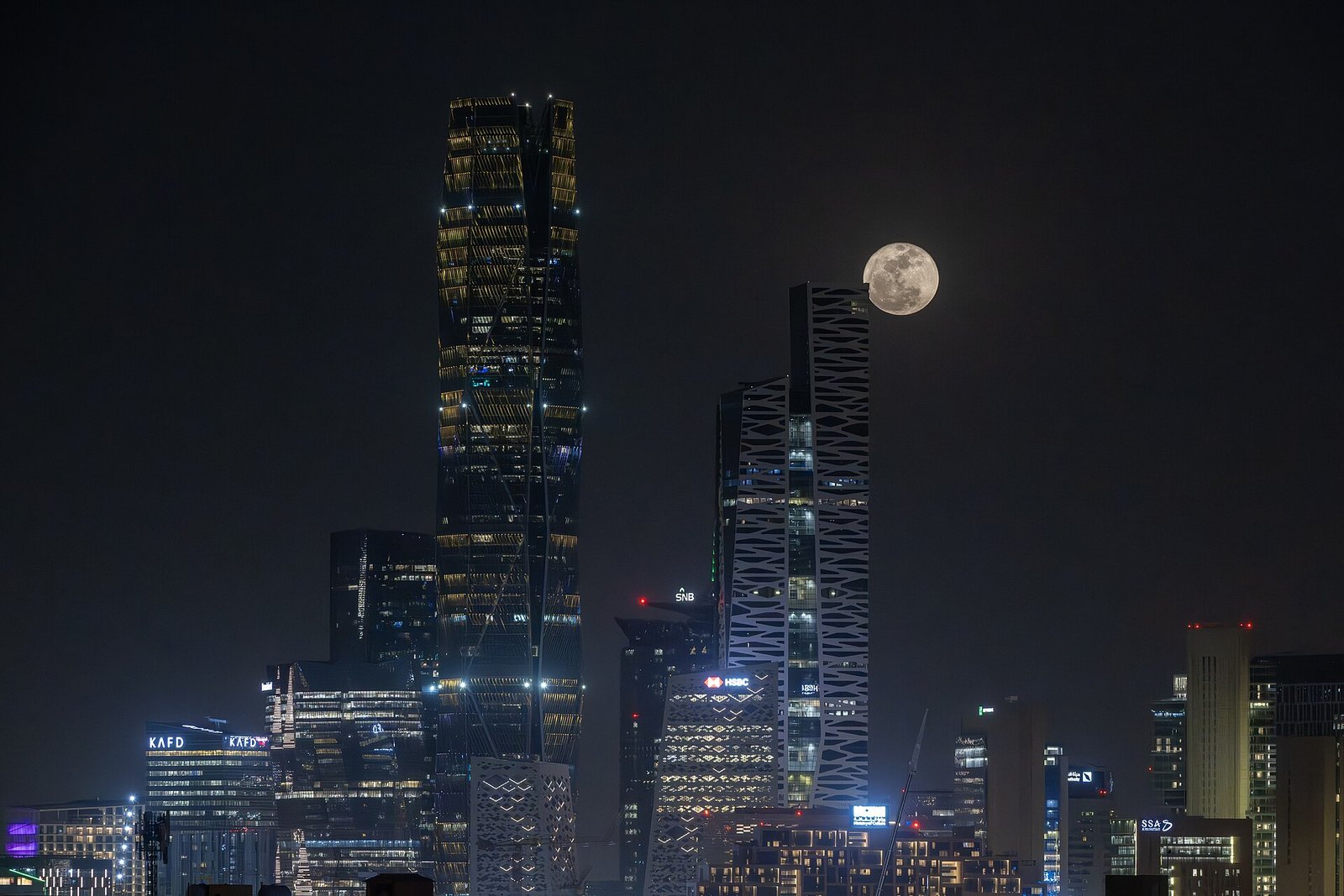 The glass towers of the King Abdullah Financial District in Riyadh lit at night beneath a full moon.