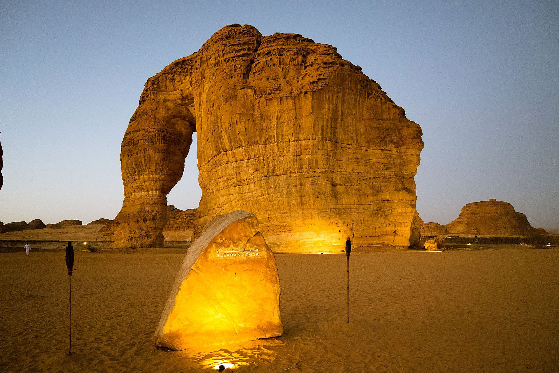Elephant Rock in AlUla glowing gold at dusk, with its lit name stone and torches in the sand