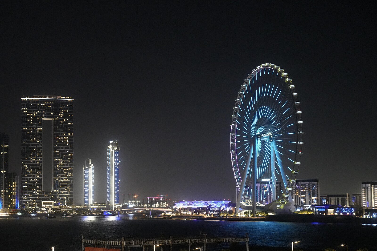 Ain Dubai glowing blue on Bluewaters Island beside the lit towers of the JBR waterfront at night.