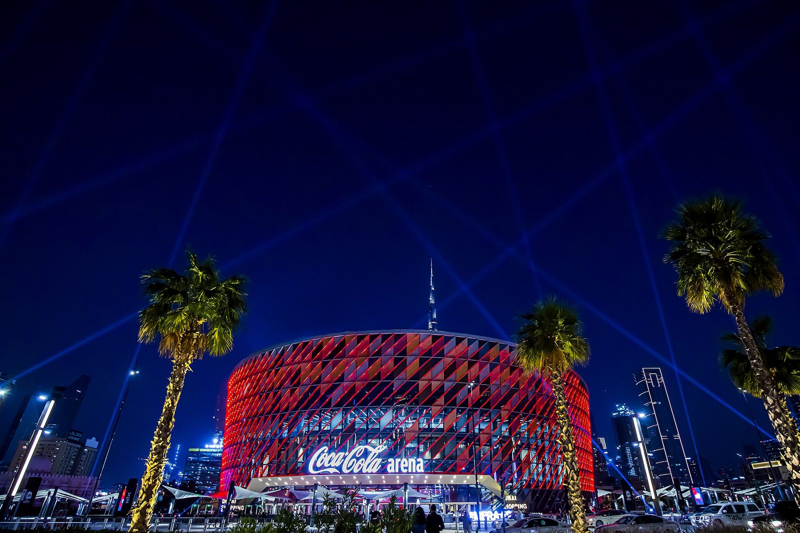 The Coca-Cola Arena in Dubai lit red at night, with searchlights and the Burj Khalifa beyond the palms
