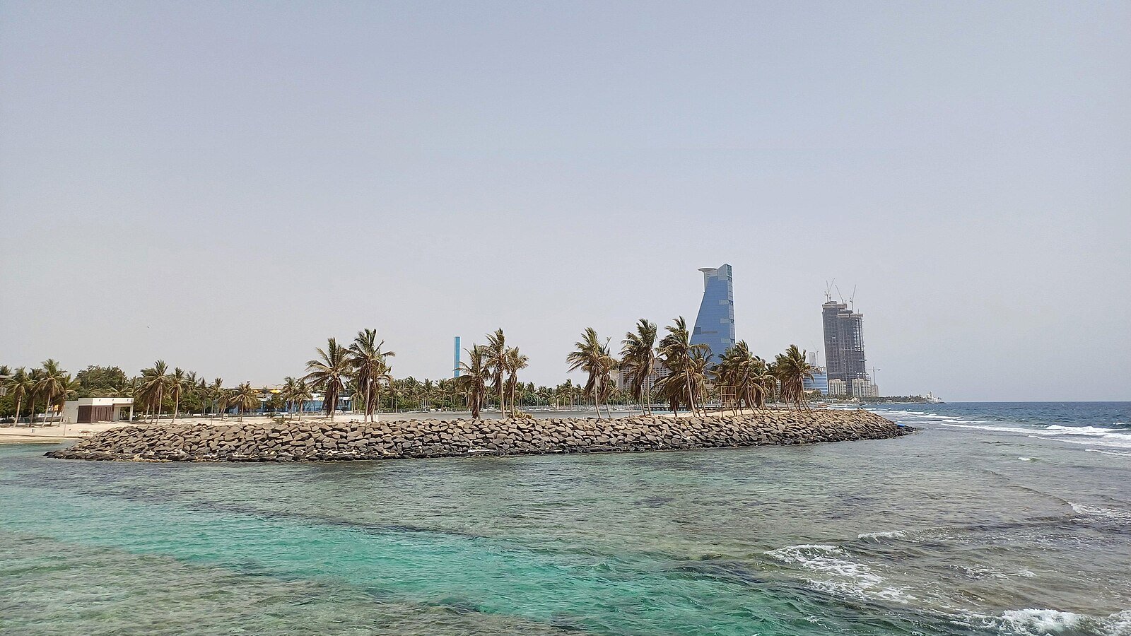Turquoise Red Sea shallows and a palm-lined breakwater along the Jeddah waterfront, with seafront towers beyond.