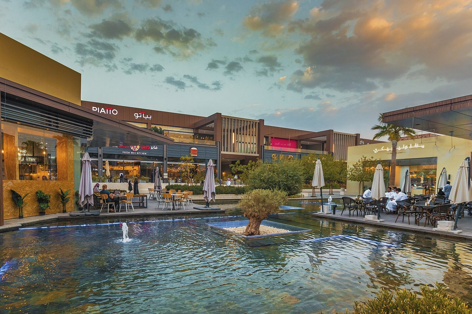 Shops, cafes and a reflecting pool with fountains at the Riyadh Front lifestyle district under an evening sky.