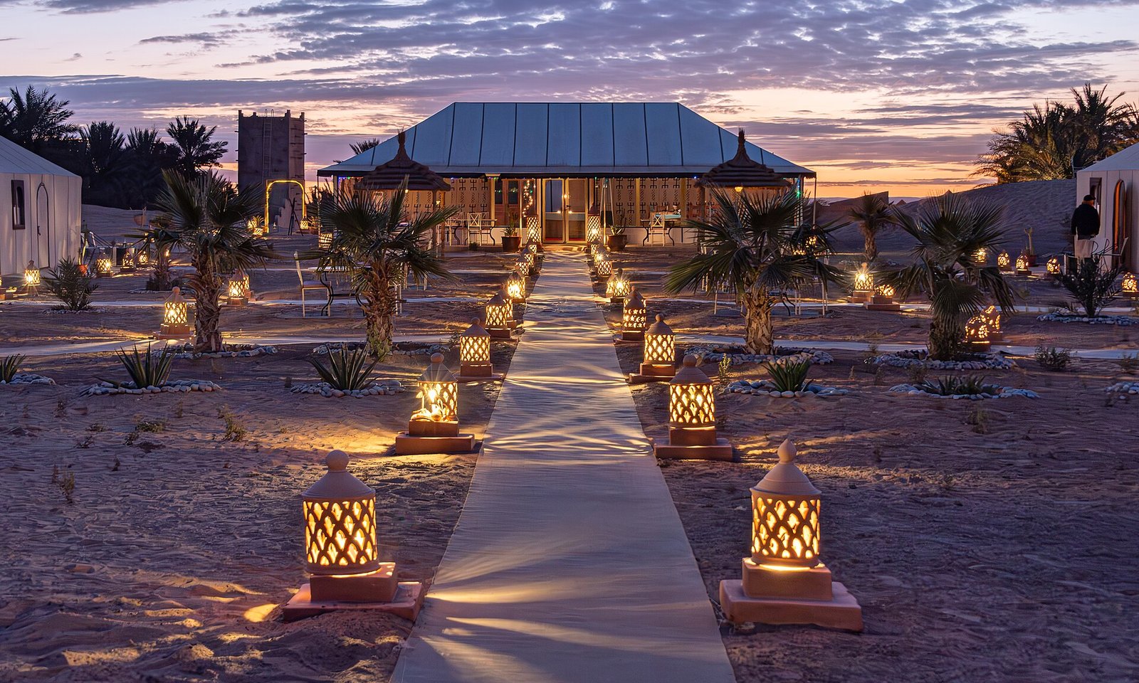 Lantern-lit paths and palms at a luxury tented camp beneath the Erg Chebbi dunes at dusk, Merzouga