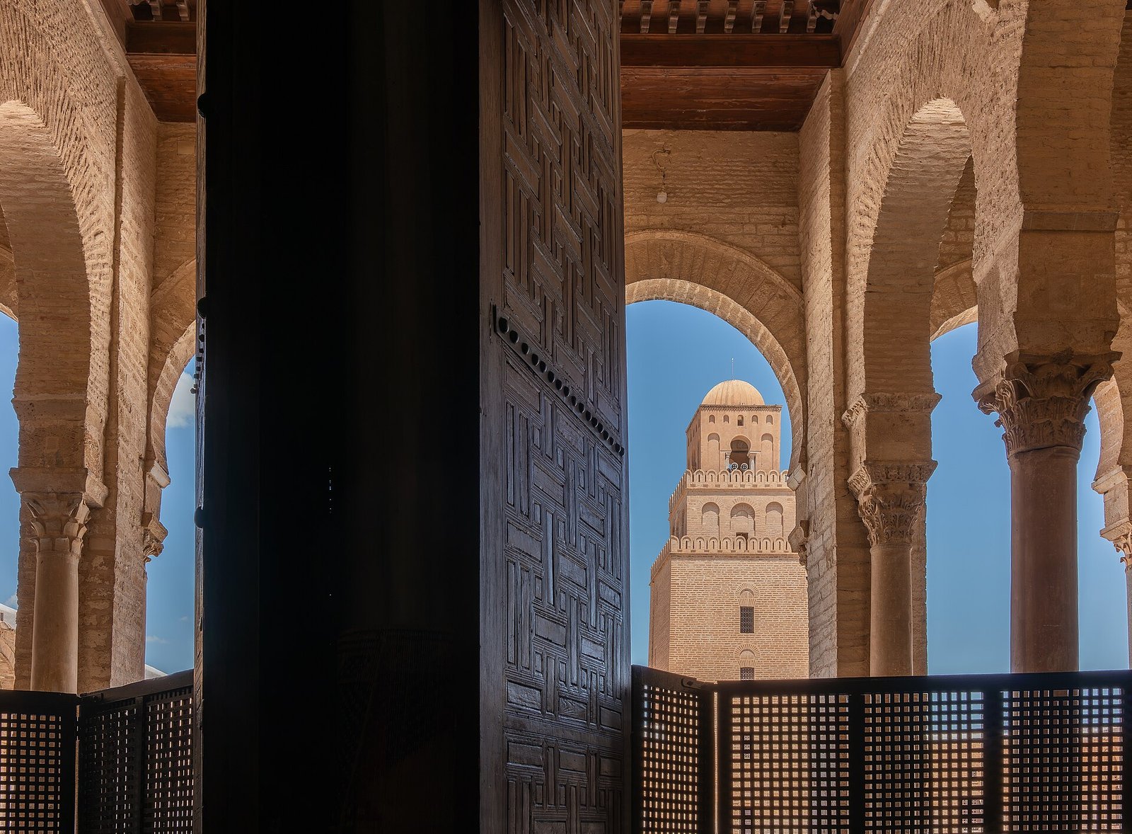 The minaret of the Great Mosque of Kairouan framed by carved horseshoe arches of the prayer-hall gallery.