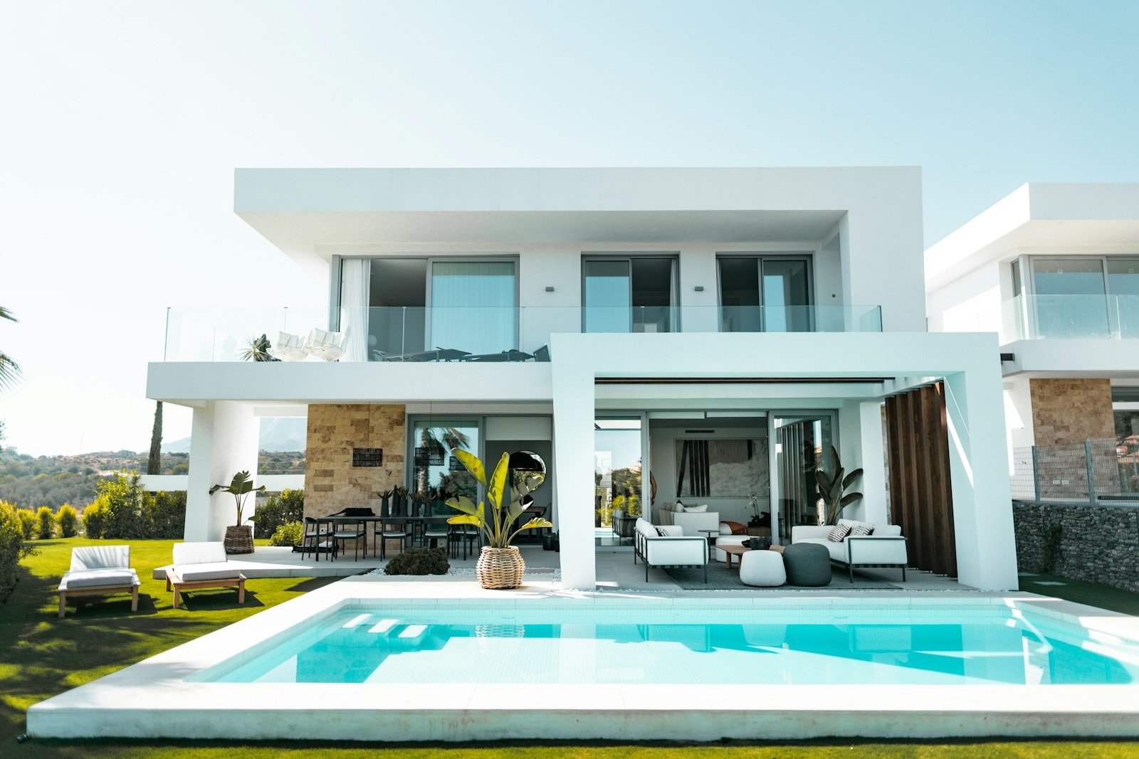 A modern white villa with an open-air lounge and sun loungers beside its private pool and lawn.