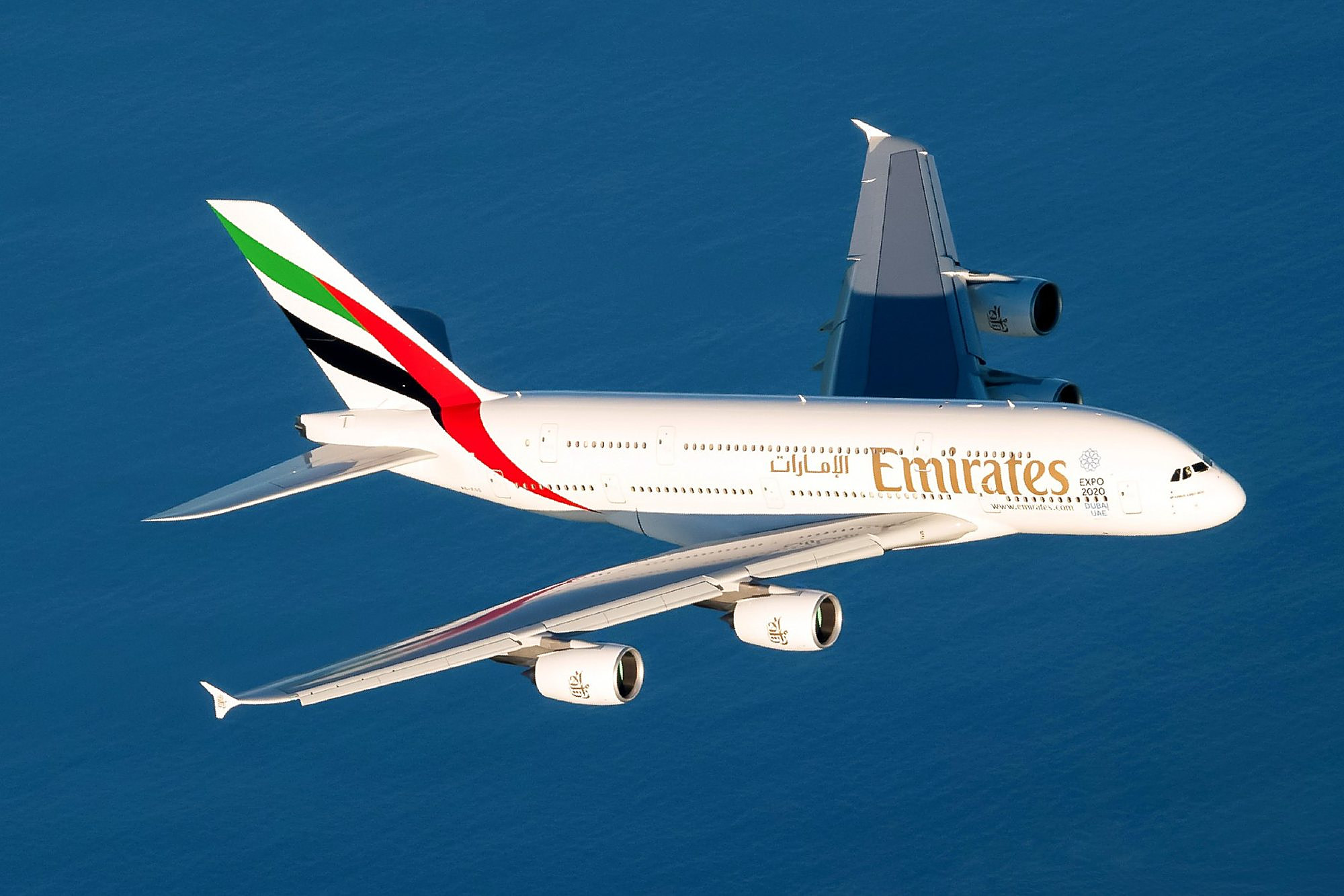 An Emirates Airbus A380 in flight seen from above, its white fuselage, gold Emirates lettering and red, green and black tail bright against deep blue sea