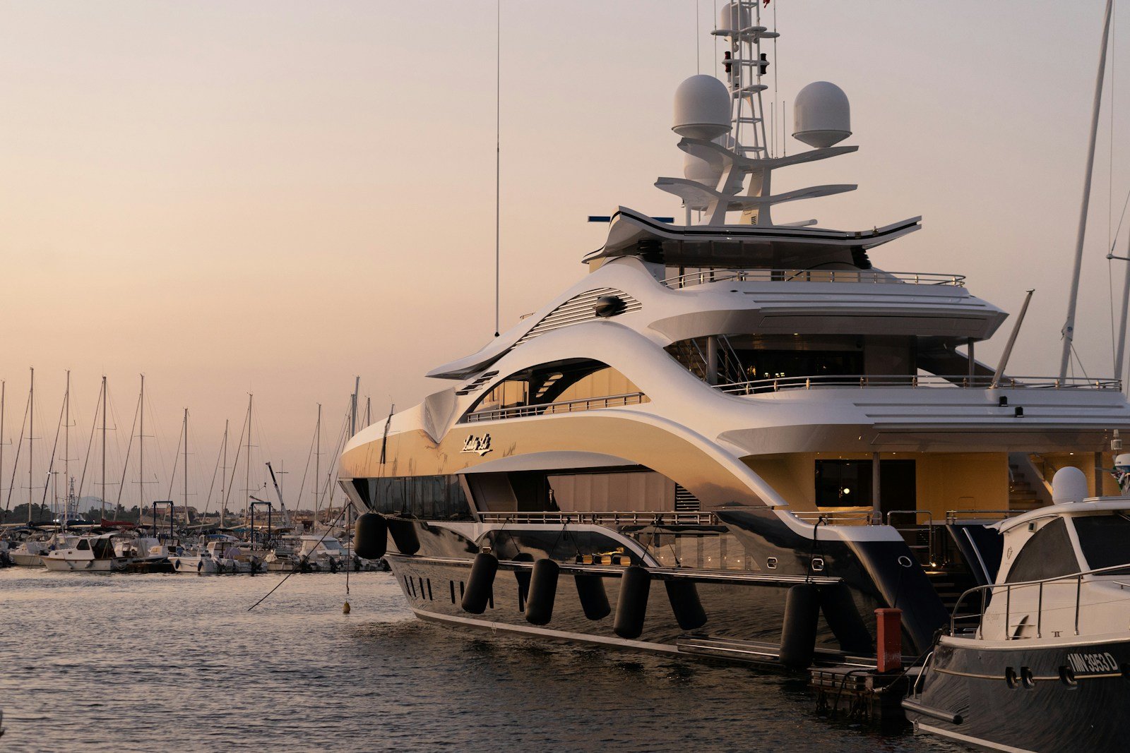 A sleek white superyacht moored at a quay in soft evening light, a forest of masts behind it.
