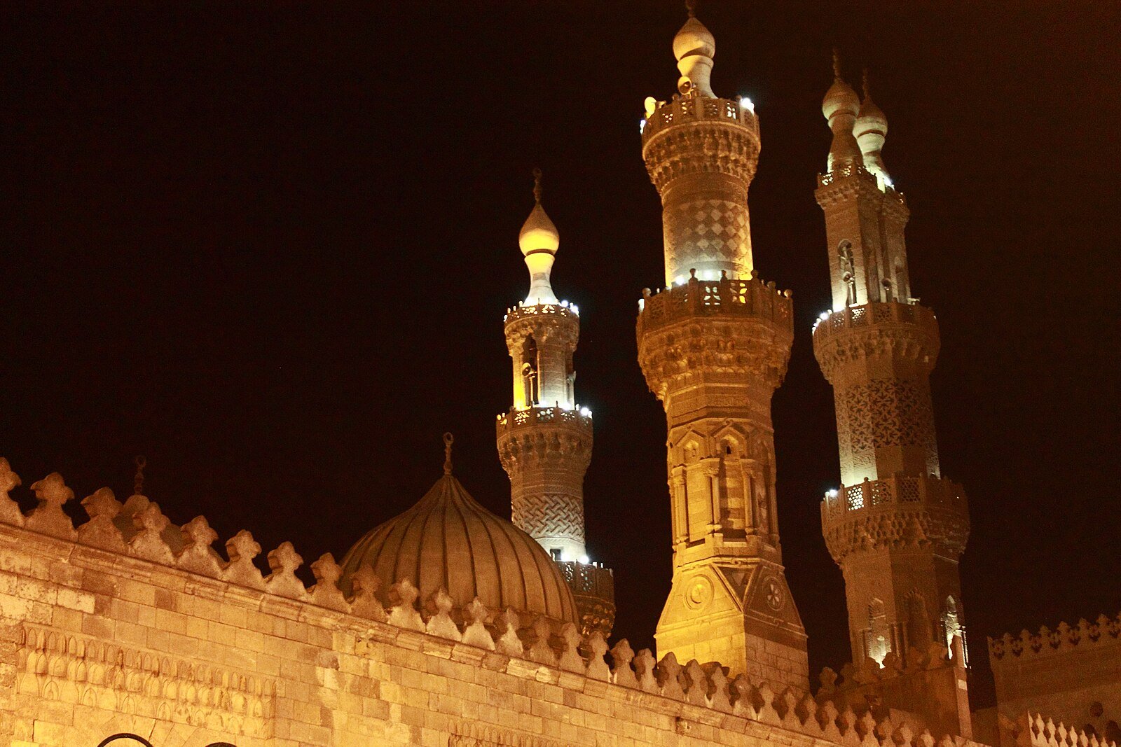 Floodlit Mamluk minarets and a ribbed stone dome rising above crenellated mosque walls in Islamic Cairo at night