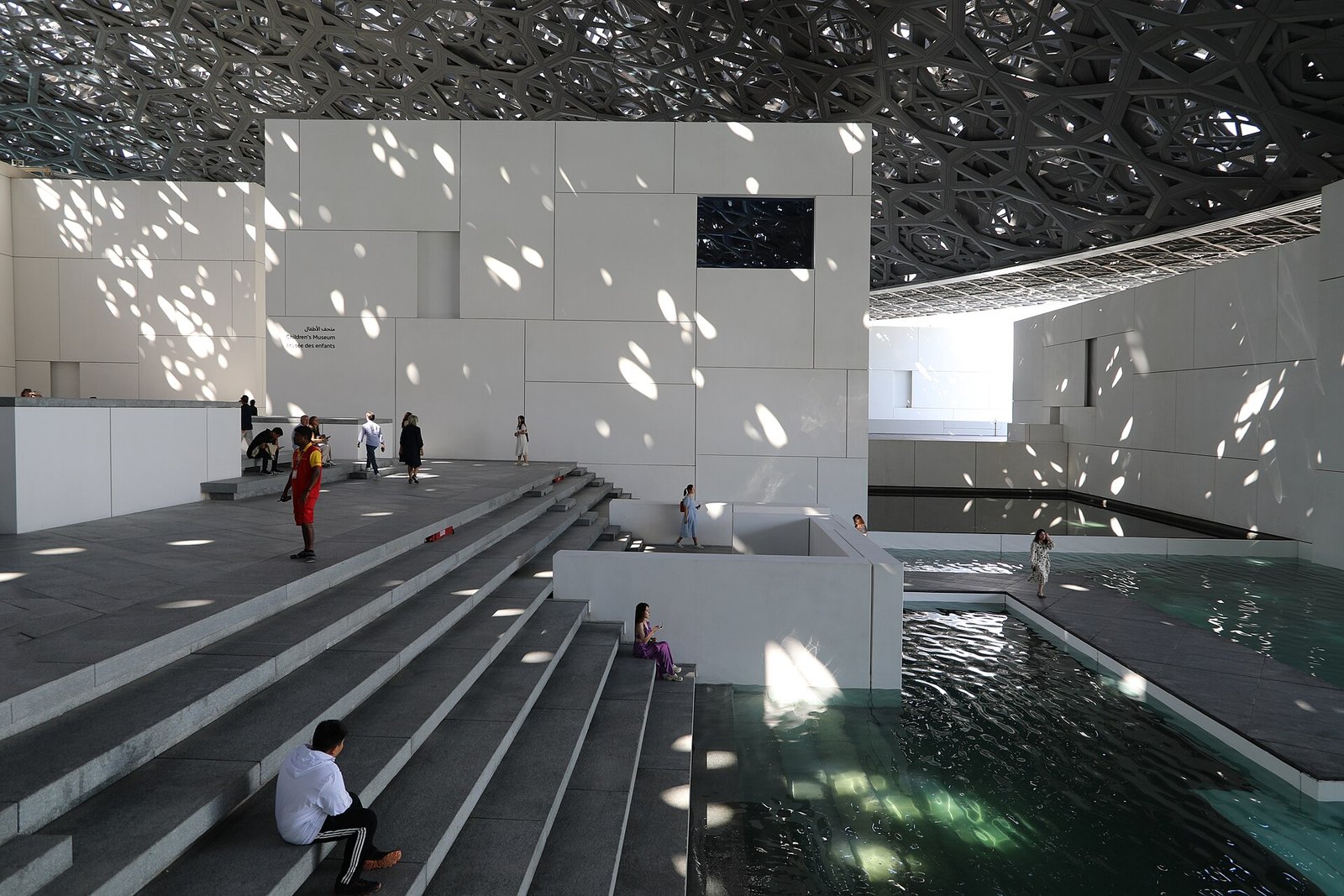 Sunlight falling through the latticed dome onto the white galleries and water of the Louvre Abu Dhabi