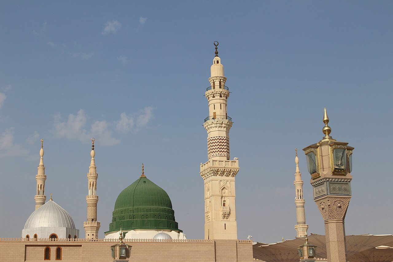 The Green Dome and white minarets of the Prophet's Mosque in Madinah against a soft blue sky.