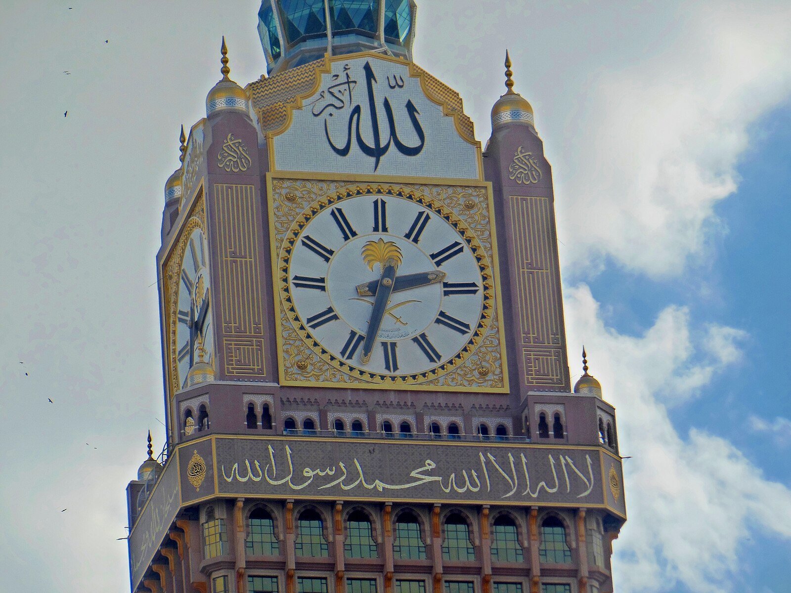 The gilded clock face and calligraphy crowning the Makkah Royal Clock Tower of Abraj Al Bait against a blue sky.